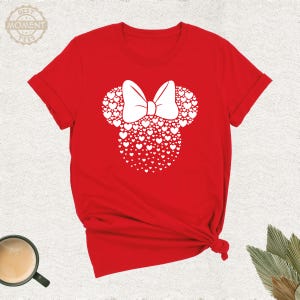 Puede incluir: Camiseta roja con un dise&ntilde;o de Minnie Mouse blanco hecho de corazones y un lazo blanco. La camiseta tiene mangas cortas y cuello redondo. El texto "BEST MOMENT TEES" est&aacute; en la esquina superior izquierda.