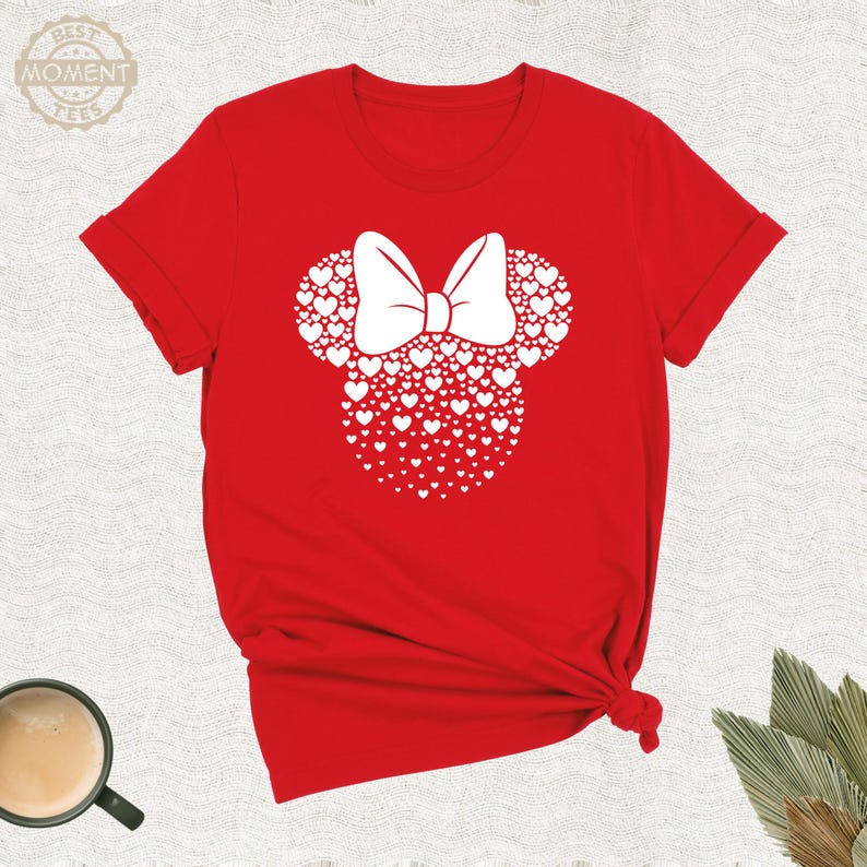 Puede incluir: Camiseta roja con un dise&ntilde;o de Minnie Mouse blanco hecho de corazones y un lazo blanco. La camiseta tiene mangas cortas y cuello redondo. El texto "BEST MOMENT TEES" est&aacute; en la esquina superior izquierda.