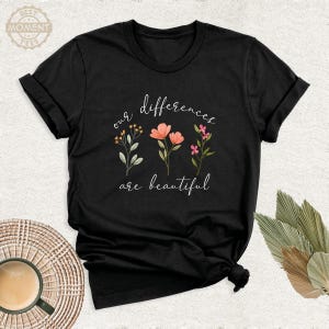 May include: A black short-sleeve t-shirt featuring a graphic of three floral stems in the center. The text "our differences are beautiful" is printed in white cursive script above and below the flowers in pink, orange, and green.