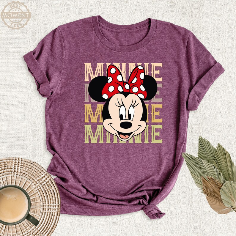 May include: A purple t-shirt with a graphic of Minnie Mouse. The graphic features Minnie Mouse's face with her signature red bow and polka dots. The text "MINNIE" is repeated three times in a vintage style font.