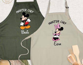 May include: Two aprons with neck loops and front pockets. The dark olive apron features Mickey Mouse with the text "MASTER CHEF" and "BRAIN". The beige apron features Minnie Mouse with the text "MASTER CHEF" and "ELENA".