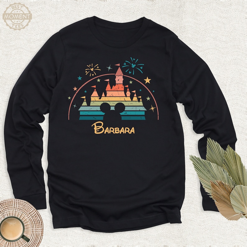 May include: A black long-sleeved shirt with a retro-style illustration of a castle with a rainbow color scheme. The castle is surrounded by stars and fireworks. The text 'BARBARA' is printed below the castle.