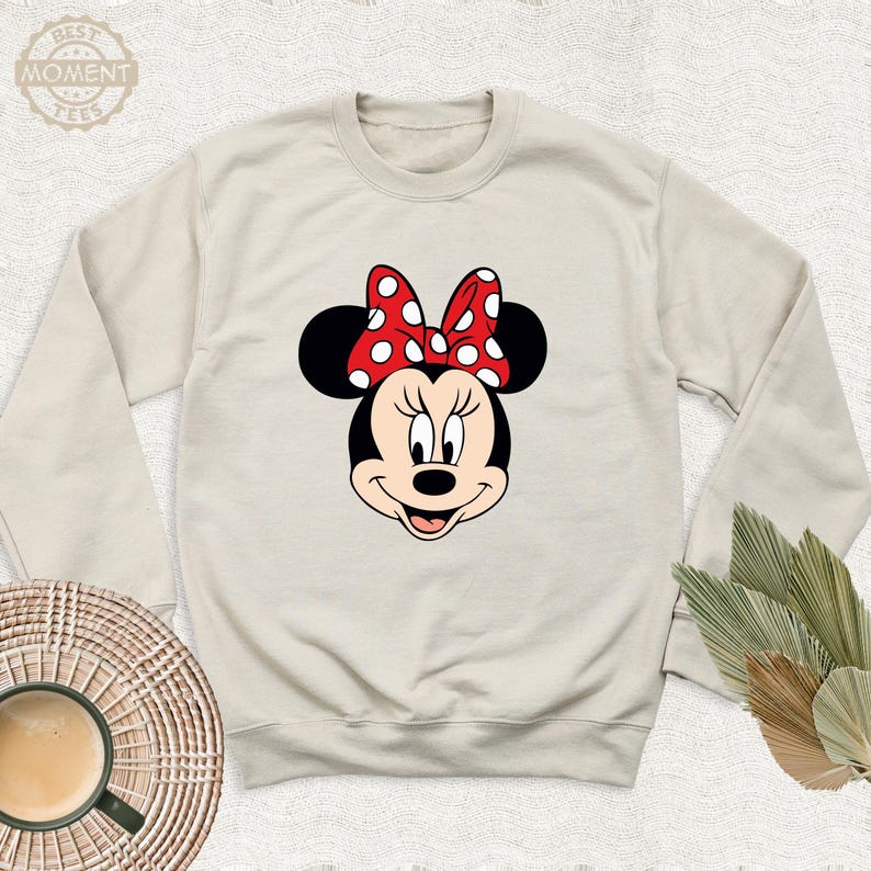 Peut inclure: Un sweat-shirt beige avec une image de dessin anim&eacute; de Minnie Mouse. Minnie Mouse porte un n&oelig;ud rouge &agrave; pois blancs.