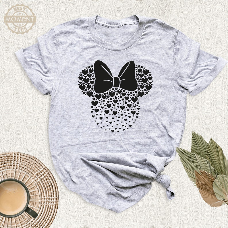 Puede incluir: Camiseta gris claro con un dise&ntilde;o de Minnie Mouse negro hecho de corazones y un lazo. La camiseta tiene mangas cortas y cuello redondo. El texto "BEST MOMENT TEES" est&aacute; en un c&iacute;rculo en la parte superior izquierda.