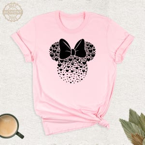 Puede incluir: Una camiseta rosa claro con un dise&ntilde;o de Minnie Mouse negro. El dise&ntilde;o incluye un lazo y orejas de rat&oacute;n hechas de corazones. El texto "BEST MOMENT TEES" est&aacute; en un c&iacute;rculo en la parte superior izquierda.