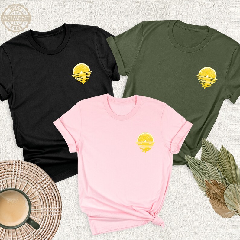 May include: Three t-shirts featuring a lemon slice sunset graphic. The shirts are black, pink and olive green.