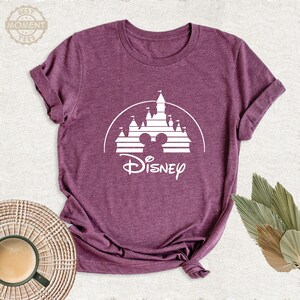 May include: A maroon t-shirt featuring a white graphic of a castle with Mickey Mouse ears and the word "Disney" in a script font. The shirt is made of a soft material and has a crew neck.