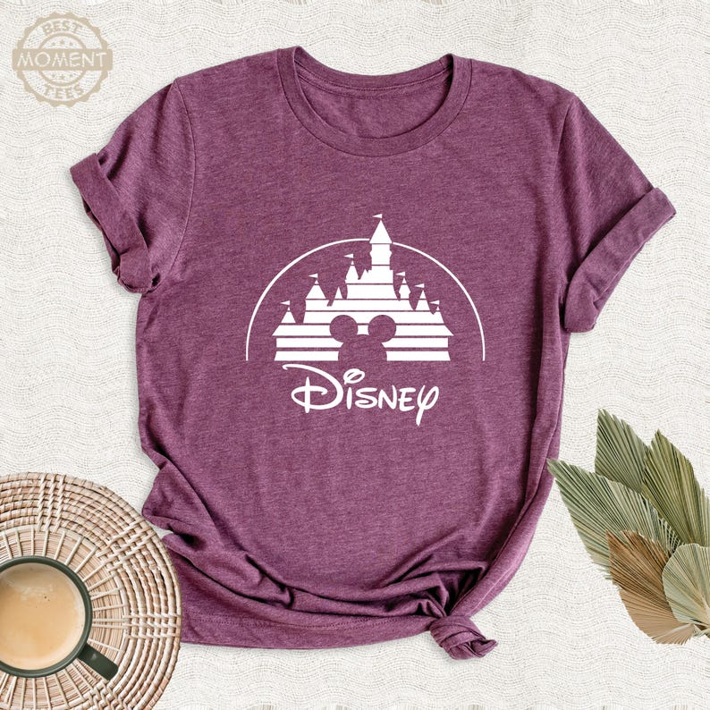 May include: A maroon t-shirt featuring a white graphic of a castle with Mickey Mouse ears and the word "Disney" in a script font. The shirt is made of a soft material and has a crew neck.