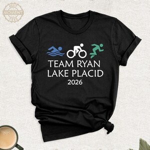 Pu&ograve; includere: T-shirt nero con grafica bianca di un nuotatore, ciclista e corridore, con la scritta "TEAM RYAN LAKE PLACID 2026". La maglietta ha maniche arrotolate e girocollo. Una tazza di caff&egrave; &egrave; visibile in basso a sinistra.
