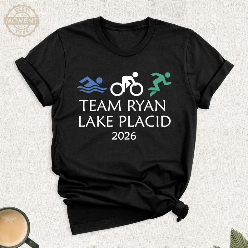 Pu&ograve; includere: T-shirt nero con grafica bianca di un nuotatore, ciclista e corridore, con la scritta "TEAM RYAN LAKE PLACID 2026". La maglietta ha maniche arrotolate e girocollo. Una tazza di caff&egrave; &egrave; visibile in basso a sinistra.