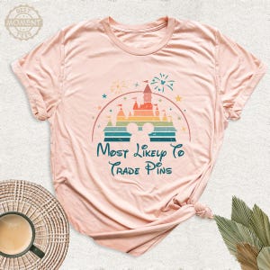 May include: A light pink t-shirt featuring a colorful graphic of a castle, fireworks, and Mickey Mouse ears. The text "Most Likely To Trade Pins" is written in teal script below the graphic.
