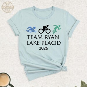 Pu&ograve; includere: Una maglietta azzurra con la scritta "TEAM RYAN LAKE PLACID 2026" in nero. Sopra il testo ci sono tre piccole icone grafiche: un nuotatore, un ciclista e un corridore. La maglietta ha le maniche arrotolate e lo scollo tondo.