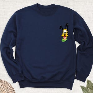 May include: Navy blue sweatshirt featuring a cartoon image of Pluto, the Disney dog, on the upper left chest. The graphic shows Pluto's face with his tongue sticking out, set against a green collar. The sweatshirt has a crew neck and long sleeves.