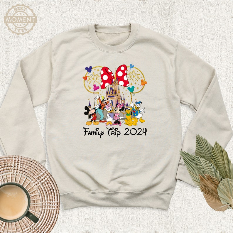 Puede incluir: Una sudadera de color beige claro con un gr&aacute;fico de Mickey Mouse y sus amigos frente al Castillo de Cenicienta. El gr&aacute;fico tambi&eacute;n incluye un lazo rojo con lunares blancos y el texto "Family Trip 2024".