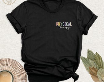 Könnte beinhalten: Ein schwarzes Kurzarm-T-Shirt mit dem Text Physical Therapy auf der linken Brustseite. Das Wort Physical ist in bunten Blockbuchstaben gehalten, während das Wort Therapy in einer weißen Schreibschrift gedruckt ist.