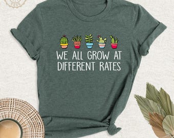 May include: A dark green short-sleeved t-shirt featuring a graphic of five colorful potted cacti and succulents above the white text that reads, WE ALL GROW AT DIFFERENT RATES. The shirt has a crew neckline and a standard fit.