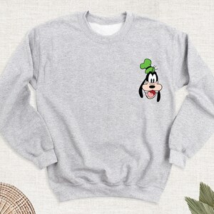 May include: A light gray sweatshirt featuring a cartoon image of Goofy. The graphic is in black, white, green, and tan. The sweatshirt has a crew neck and long sleeves. The design is on the left chest area.