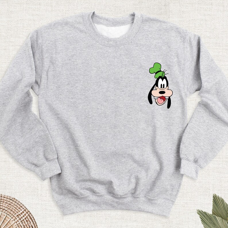 May include: A light gray sweatshirt featuring a cartoon image of Goofy. The graphic is in black, white, green, and tan. The sweatshirt has a crew neck and long sleeves. The design is on the left chest area.