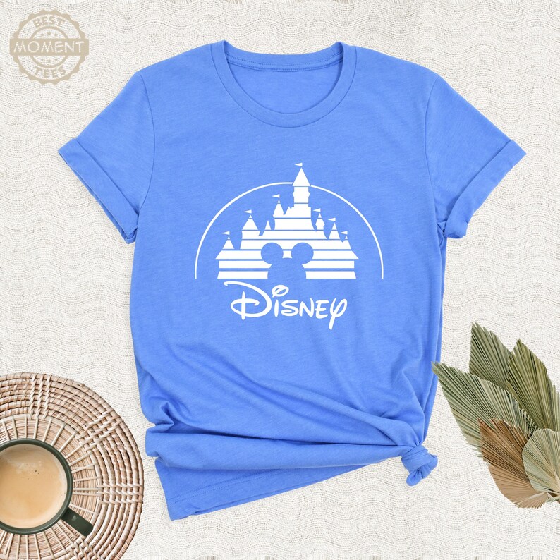 May include: A light blue t-shirt featuring a white graphic of a castle, Mickey Mouse ears, and the word "Disney". The shirt has short sleeves and a crew neck. The text "Best Moment Tees" is at the top left.