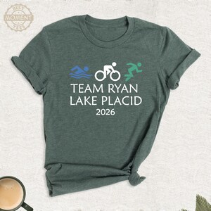 Pu&ograve; includere: T-shirt verde con la scritta "TEAM RYAN LAKE PLACID 2026" e illustrazioni di nuoto, ciclismo e corsa. Il design presenta grafiche blu, bianche e verdi, per gli appassionati di sport.