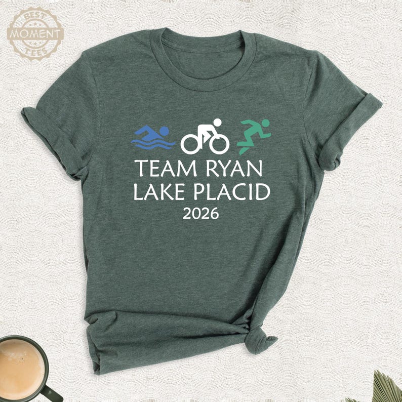 Pu&ograve; includere: T-shirt verde con la scritta "TEAM RYAN LAKE PLACID 2026" e illustrazioni di nuoto, ciclismo e corsa. Il design presenta grafiche blu, bianche e verdi, per gli appassionati di sport.