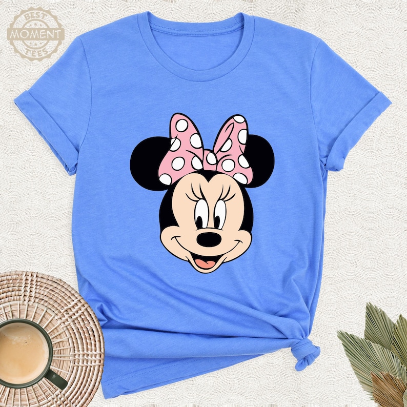 Puede incluir: Una camiseta azul claro con un gr&aacute;fico de Minnie Mouse. El gr&aacute;fico muestra la cara de Minnie con un lazo rosa con lunares blancos. La camiseta tiene mangas enrolladas y el texto "BEST MOMENT TEES" es visible.