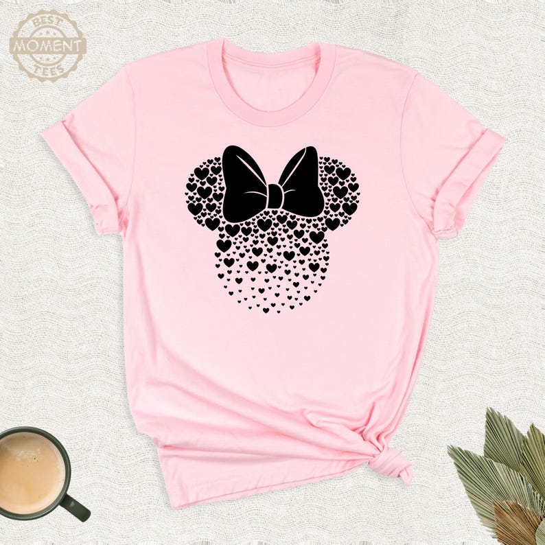 Puede incluir: Una camiseta rosa claro con un dise&ntilde;o de Minnie Mouse negro. El dise&ntilde;o incluye un lazo y orejas de rat&oacute;n hechas de corazones. El texto "BEST MOMENT TEES" est&aacute; en un c&iacute;rculo en la parte superior izquierda.