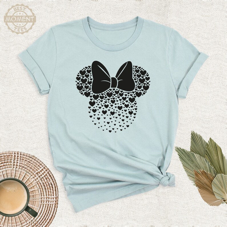 Puede incluir: Camiseta azul claro con un dise&ntilde;o de Minnie Mouse negro hecho de corazones y un lazo. La camiseta tiene mangas enrolladas y un dobladillo anudado. El texto "BEST MOMENT TEES" est&aacute; impreso en la parte superior izquierda.