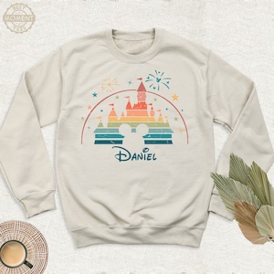 May include: A light beige sweatshirt with a retro-style illustration of a castle with a rainbow color scheme. The castle is surrounded by stars and fireworks. The text "Daniel" is printed below the castle.