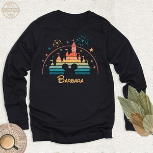 May include: A black long-sleeve shirt with a retro-style graphic of a rainbow-colored Cinderella's castle with fireworks and stars. The graphic also includes a Mickey Mouse silhouette and the name "BARBARA" below the castle.
