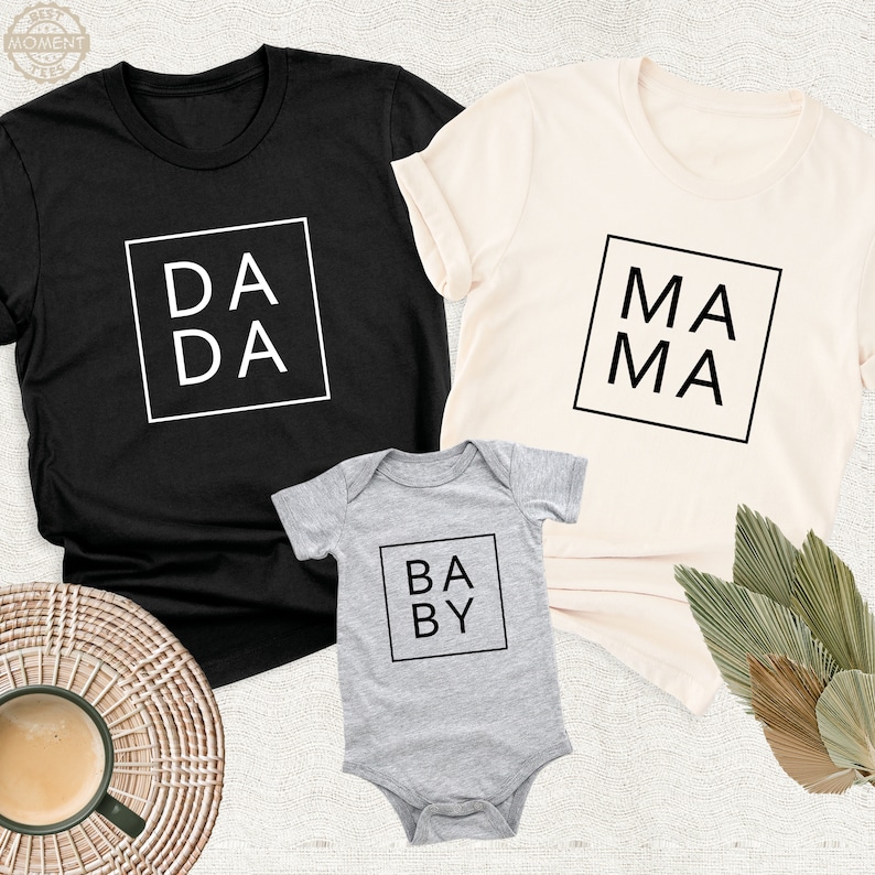 Peut inclure: Un ensemble de trois t-shirts avec les mots "DA DA", "MA MA" et "BABY" imprim&eacute;s en noir sur fond blanc. Les t-shirts sont noir, cr&egrave;me et gris.