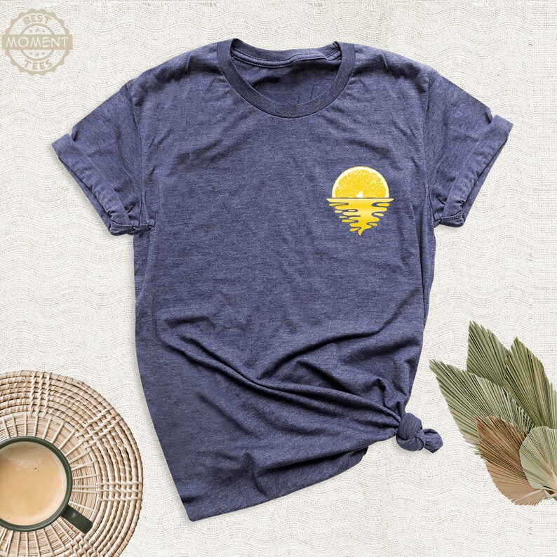 May include: A navy blue t-shirt featuring a yellow lemon slice graphic. The lemon slice is depicted as a sunset over a body of water.