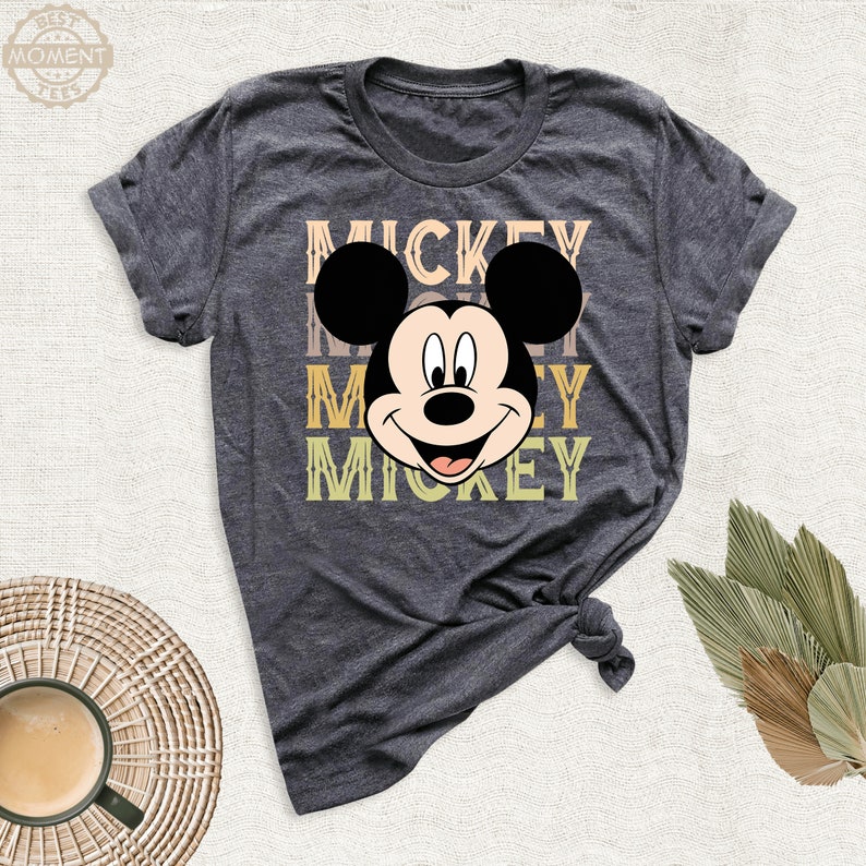 May include: A dark gray t-shirt with a retro-style graphic of Mickey Mouse's face and the word "Mickey" repeated three times in different colors.