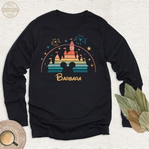 May include: A black long-sleeve shirt with a retro-style graphic of a rainbow-colored Cinderella's castle with fireworks and stars. The graphic also includes a Mickey Mouse silhouette and the name "BARBARA" below the castle.