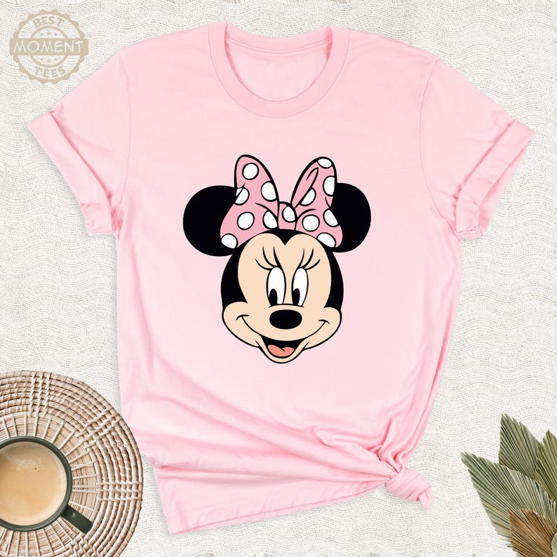 Puede incluir: Camiseta rosa con un dise&ntilde;o de Minnie Mouse. El dise&ntilde;o muestra la cara de Minnie, con orejas negras y un lazo rosa con lunares blancos. La camiseta tiene mangas cortas y cuello redondo.