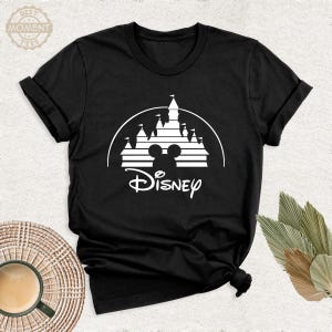 May include: Black t-shirt featuring a white graphic of a castle silhouette, Mickey Mouse ears, and the word "Disney" in a script font. The shirt has short sleeves and a crew neck. The image is taken from a slightly elevated angle.