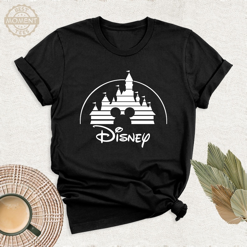 May include: Black t-shirt featuring a white graphic of a castle silhouette, Mickey Mouse ears, and the word "Disney" in a script font. The shirt has short sleeves and a crew neck. The image is taken from a slightly elevated angle.