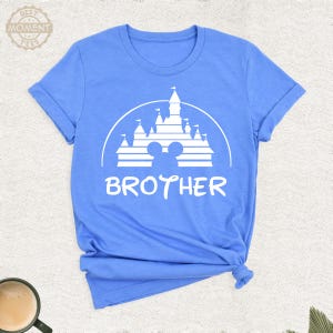 Puede incluir: Camiseta azul claro con un gr&aacute;fico blanco de un castillo, orejas de Mickey Mouse y la palabra "BROTHER". La camiseta tiene mangas cortas y cuello redondo.