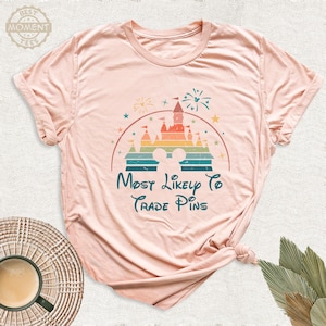 May include: A light pink t-shirt featuring a colorful graphic of a castle, fireworks, and Mickey Mouse ears. The text "Most Likely To Trade Pins" is written in teal script below the graphic.
