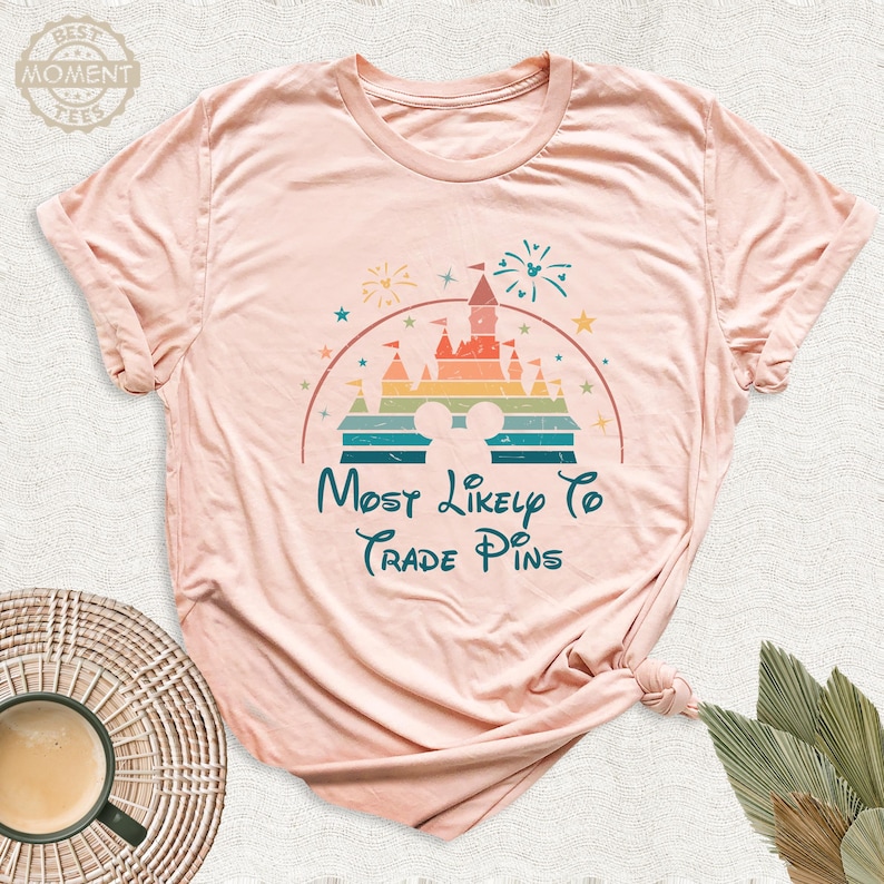 May include: A light pink t-shirt featuring a colorful graphic of a castle, fireworks, and Mickey Mouse ears. The text "Most Likely To Trade Pins" is written in teal script below the graphic.