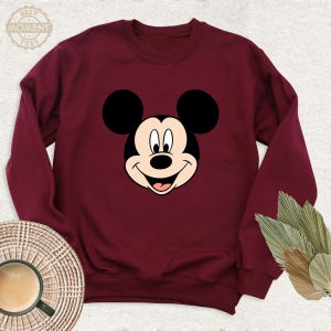 May include: A maroon sweatshirt featuring a cartoon image of Mickey Mouse's face. Mickey Mouse is smiling with his iconic black ears and a black nose.