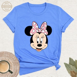 Puede incluir: Camiseta azul claro con un gr&aacute;fico de Minnie Mouse. El gr&aacute;fico muestra la cara de Minnie con un lazo rosa con lunares blancos. La camiseta tiene mangas enrolladas y el texto "BEST MOMENT TEES" es visible.