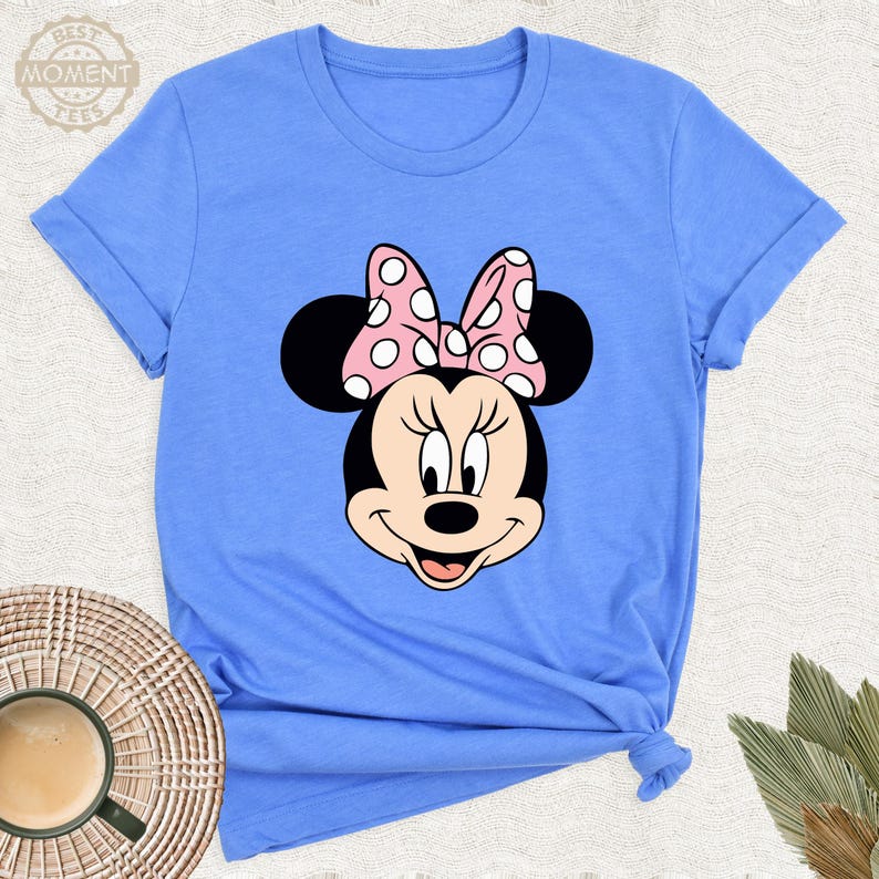 Puede incluir: Camiseta azul claro con un gr&aacute;fico de Minnie Mouse. El gr&aacute;fico muestra la cara de Minnie con un lazo rosa con lunares blancos. La camiseta tiene mangas enrolladas y el texto "BEST MOMENT TEES" es visible.