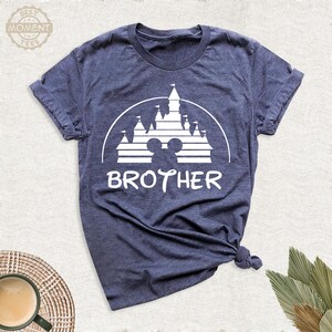Puede incluir: Camiseta azul marino con un gr&aacute;fico blanco de un castillo, orejas de Mickey Mouse y la palabra "BROTHER". La camiseta est&aacute; hecha de un material suave y tiene mangas cortas. El dise&ntilde;o est&aacute; impreso en la parte delantera.