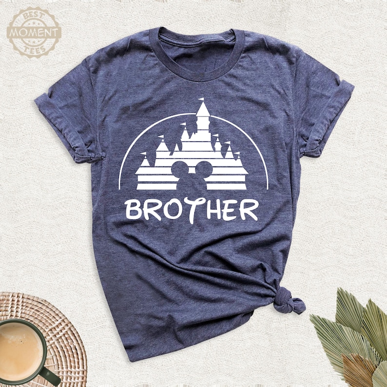 Puede incluir: Camiseta azul marino con un gr&aacute;fico blanco de un castillo, orejas de Mickey Mouse y la palabra "BROTHER". La camiseta est&aacute; hecha de un material suave y tiene mangas cortas. El dise&ntilde;o est&aacute; impreso en la parte delantera.