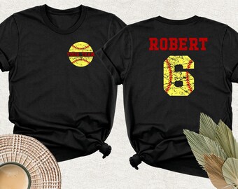 May include: A black t-shirt featuring a yellow softball graphic with red stitching on the front left chest. The back displays the name ROBERT in red text above a large yellow softball-textured number 6 with red stitching.