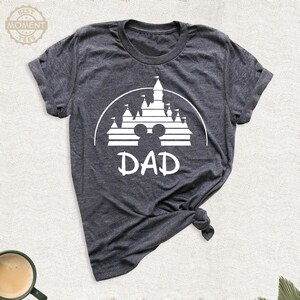 Puede incluir: Camiseta gris oscuro con un gr&aacute;fico blanco de un castillo, silueta de Mickey Mouse y la palabra "DAD". La camiseta est&aacute; hecha de un material suave y tiene cuello redondo. La camiseta est&aacute; anudada en la parte inferior.