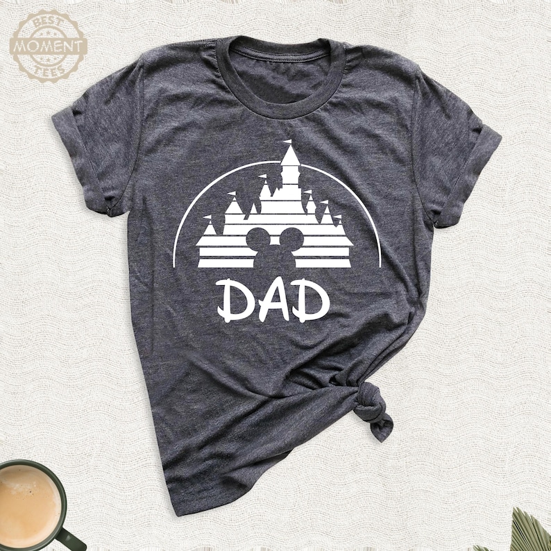 Puede incluir: Camiseta gris oscuro con un gr&aacute;fico blanco de un castillo, silueta de Mickey Mouse y la palabra "DAD". La camiseta est&aacute; hecha de un material suave y tiene cuello redondo. La camiseta est&aacute; anudada en la parte inferior.
