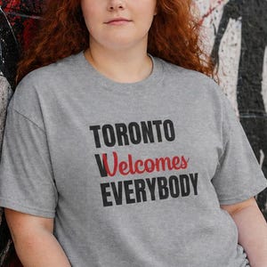 以下が含まれることがあります： 「TORONTO Welcomes EVERYBODY」と黒と赤で書かれたグレーのTシャツ。青いジーンズを履いた人が、サッカーボールの壁画と「TOGETHER We Play」と書かれた落書きのある壁に寄りかかっています。