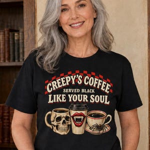 Puede incluir: Una camiseta negra de manga corta con un gráfico de una taza con forma de calavera, un vaso de café con colmillos de vampiro y una taza de té con una araña. El texto dice: CREEPY'S COFFEE SERVED BLACK LIKE YOUR SOUL. Patrón de cuadros rojo y negro.